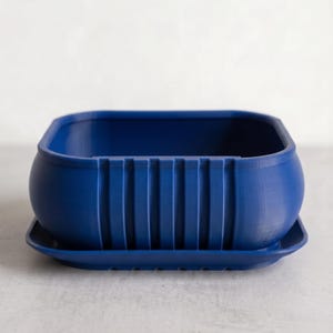 May include: A dark blue rectangular plant pot with a matching tray. The pot has a rounded body and vertical ribbed detailing on the front. The tray is rectangular with a slightly raised edge. The pot and tray are made of a solid, matte material.