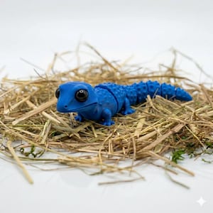 Könnte beinhalten: Ein leuchtend blaues, bewegliches Echsenspielzeug mit großen schwarzen Augen, das auf einem Heubett liegt. Der segmentierte Körper und die strukturierte Haut der Eidechse sind deutlich sichtbar. Diese skurrile Reptilienfigur ist ein einzigartiger Dekorationsartikel.