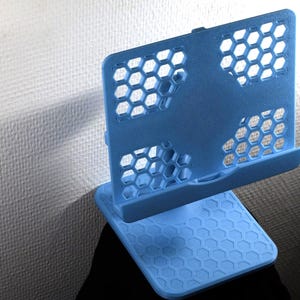 Könnte beinhalten: Ein hellblauer Tablet- oder Handyhalter mit einem geometrischen Wabenmuster auf der hinteren Stützplatte und einem passenden hexagonal strukturierten Sockel. Der Halter verfügt über eine horizontale Ablage an der Unterseite für das Gerät.