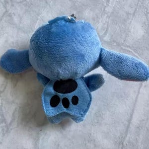Puede incluir: Un llavero de peluche azul que representa un personaje con orejas grandes y un dise&ntilde;o de huella de pata en el vientre. El llavero tiene un anillo de metal plateado adjunto en la parte superior. El juguete est&aacute; hecho de material suave y difuso.