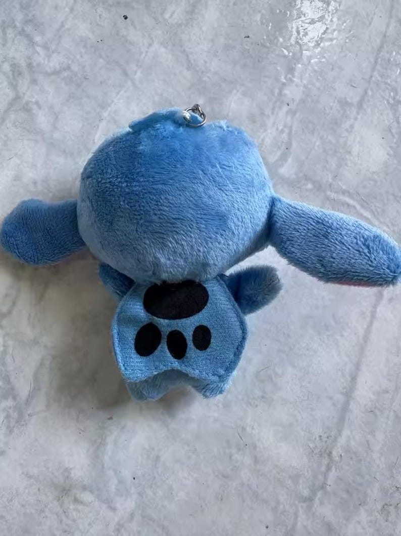 Puede incluir: Un llavero de peluche azul que representa un personaje con orejas grandes y un dise&ntilde;o de huella de pata en el vientre. El llavero tiene un anillo de metal plateado adjunto en la parte superior. El juguete est&aacute; hecho de material suave y difuso.