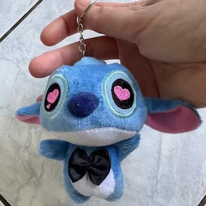 Puede incluir: Un llavero de peluche azul con un personaje de dibujos animados con ojos grandes, pupilas en forma de coraz&oacute;n rosa y una pajarita negra. El llavero est&aacute; sujeto a un anillo de metal plateado.