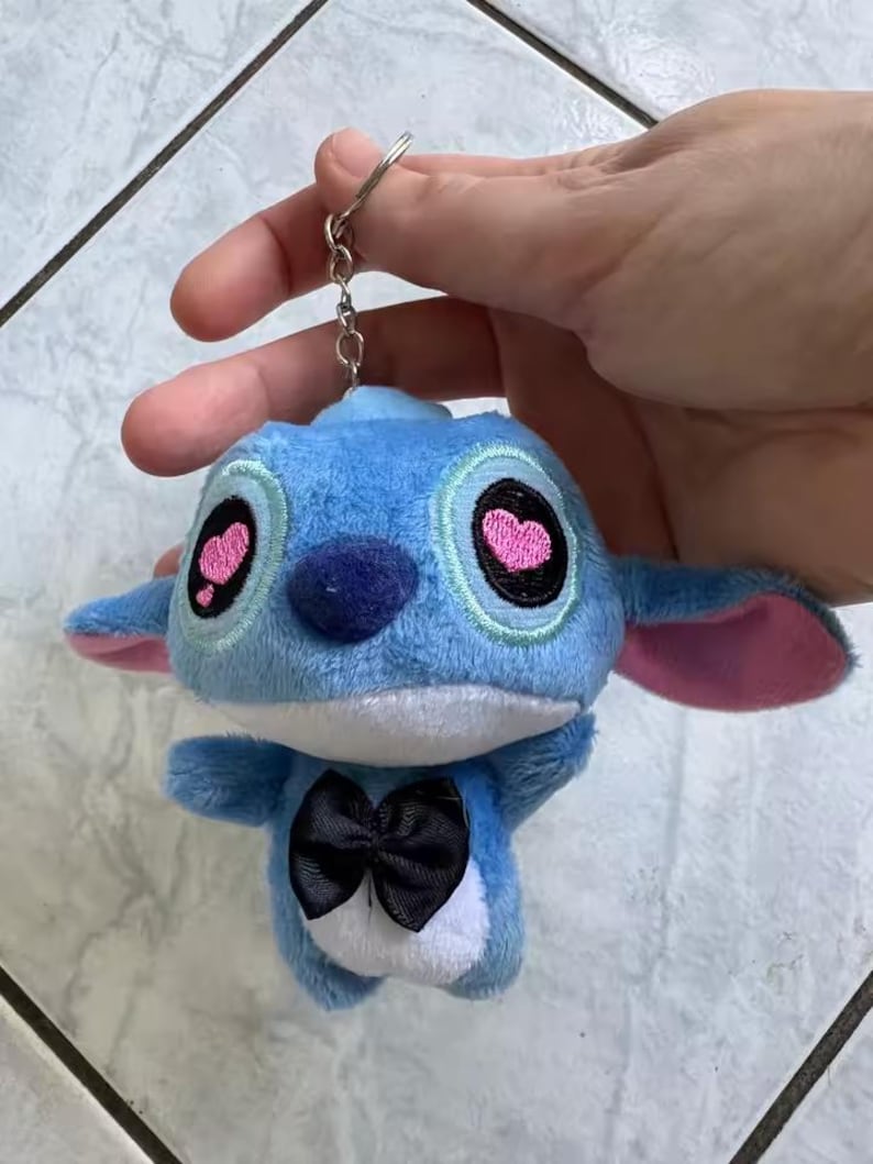 Puede incluir: Un llavero de peluche azul con un personaje de dibujos animados con ojos grandes, pupilas en forma de coraz&oacute;n rosa y una pajarita negra. El llavero est&aacute; sujeto a un anillo de metal plateado.