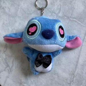 Puede incluir: Llavero de peluche azul con un personaje de orejas grandes, detalles rosas y una pajarita negra. El llavero tiene un anillo de metal plateado para sujetarlo. Los ojos del personaje son negros con pupilas rosas en forma de coraz&oacute;n.
