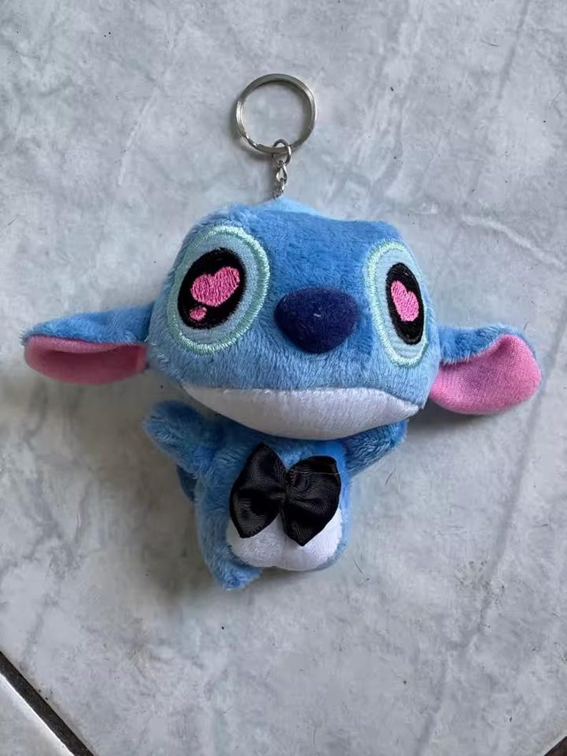 Puede incluir: Llavero de peluche azul con un personaje de orejas grandes, detalles rosas y una pajarita negra. El llavero tiene un anillo de metal plateado para sujetarlo. Los ojos del personaje son negros con pupilas rosas en forma de coraz&oacute;n.