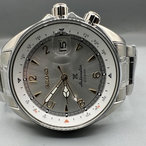 May include: A silver-toned Seiko automatic wristwatch with a brushed metallic dial, featuring gold-colored Arabic numerals at 2, 4, 6, 8, 10, and 12 o'clock, a date window at 3 o'clock, a rotating compass bezel, and a stainless steel link bracelet.