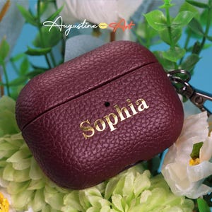 Könnte beinhalten: Burgunderfarbenes Leder-AirPod-Case mit dem Namen "Sophia" in goldfarbenen Buchstaben. Das Case hat eine strukturierte Oberfläche und einen silberfarbenen Verschluss. Der Hintergrund zeigt grünes Laub und weiße Blüten.
