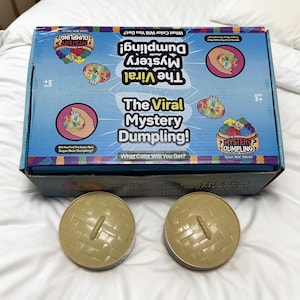 Può includere: Una scatola blu con la scritta "The Viral Mystery Dumpling!" e illustrazioni colorate di ravioli. Due contenitori rotondi beige con una trama intrecciata sono davanti alla scatola. La scatola dice anche "What Color Will You Get?" e "Sugar Bear Edition."
