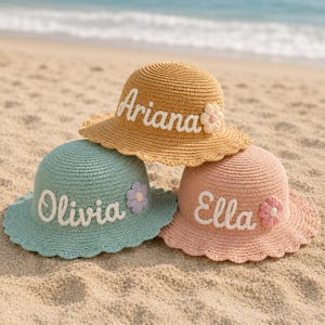 Puede incluir: Tres sombreros de sol de paja en amarillo, verde azulado y rosa, cada uno con un nombre bordado en blanco: "Ariana", "Olivia" y "Ella". Cada sombrero tiene una flor decorativa. Los sombreros están en una playa de arena.