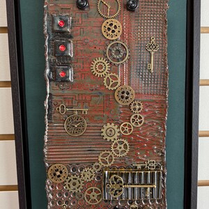 May include: A framed mixed-media artwork with a steampunk theme. The piece features a textured, reddish-brown background adorned with brass gears, keys, and clock faces. Red, circular accents and black hardware add contrast.
