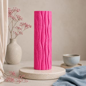 May include: A tall, cylindrical, hot pink vase with a textured, vertical, wavy pattern. The vase is displayed on a round, light-colored pedestal. A small, light-colored vase with pink flowers is in the background.