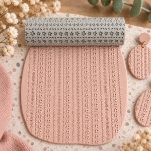 May include: A grey cylindrical rolling pin with a repeating floral pattern and a flat sheet of light pink clay featuring the same embossed flower and leaf design, accompanied by two matching oval-shaped clay earrings with gold-tone hardware.