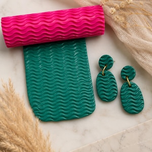May include: A hot pink textured rolling pin rests atop a teal green sheet with a wavy pattern. Two teal green earrings with gold-colored hoops are next to the sheet. The earrings and sheet have the same wavy pattern.