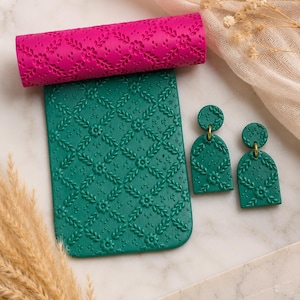 May include: A vibrant pink textured rolling pin sits above a teal green rectangular slab, both featuring a floral design. Two matching teal green earrings with gold hoops are also visible. The items are arranged on a white surface.