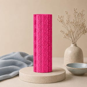 May include: A bright pink vase with a textured, vertical design. The cylindrical vase is displayed on a circular, pale-coloured plinth. A blue bowl and a small vase with dried flowers are in the background.