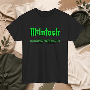Könnte beinhalten: Schwarzes T-Shirt mit dem Wort "McIntosh" in grüner gotischer Schrift. Darunter steht der Satz "LEGENDARY PERFORMANCE" ebenfalls in Grün. Im Hintergrund sind weitere Kleidungsstücke wie Kapuzenpullover zu sehen.