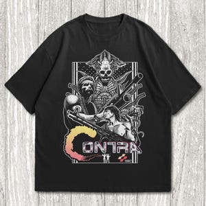 K&ouml;nnte beinhalten: Ein schwarzes Kurzarm-T-Shirt mit einem detaillierten wei&szlig;en Grafikdruck von zwei muskul&ouml;sen Soldaten, die gro&szlig;e Gewehre halten. &Uuml;ber ihnen befindet sich eine totenkopfartige Kreatur. Das Wort CONTRA ist in einem Farbverlauf aufgedruckt.