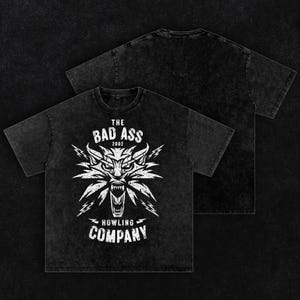 Peut inclure: Un t-shirt noir d&eacute;lav&eacute; &agrave; l'acide avec un motif blanc repr&eacute;sentant une t&ecirc;te de loup stylis&eacute;e et des &eacute;clairs. Le texte indique THE BAD ASS 2002 HOWLING COMPANY. Le motif pr&eacute;sente un aspect vieilli et us&eacute; sur le devant, le dos est uni.