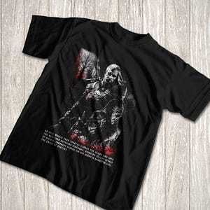 May include: A black short-sleeve t-shirt featuring a graphic illustration of a warrior with a sword. The design includes red and white accents and text that reads: An epic world tour travels across the globe and into the war-torn northern kingdoms.