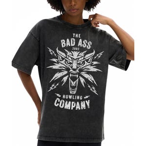 Peut inclure: Un t-shirt gris anthracite d&eacute;lav&eacute; &agrave; l'acide avec une impression graphique blanche repr&eacute;sentant une t&ecirc;te de loup stylis&eacute;e gueule ouverte, entour&eacute;e d'&eacute;clairs. Le texte indique THE BAD ASS 2002 HOWLING COMPANY dans une police blanche vieillie.