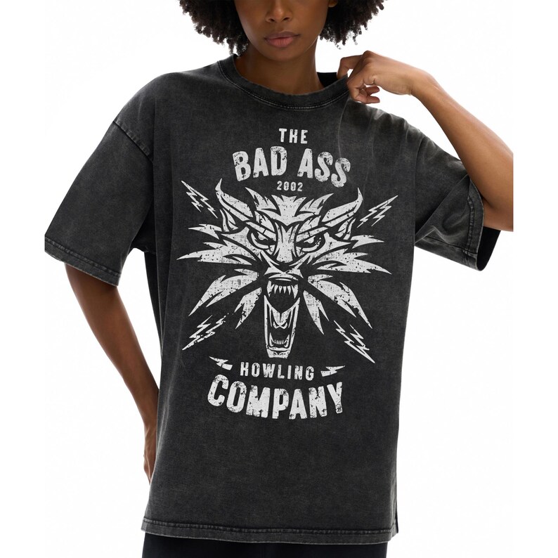 Peut inclure: Un t-shirt gris anthracite d&eacute;lav&eacute; &agrave; l'acide avec une impression graphique blanche repr&eacute;sentant une t&ecirc;te de loup stylis&eacute;e gueule ouverte, entour&eacute;e d'&eacute;clairs. Le texte indique THE BAD ASS 2002 HOWLING COMPANY dans une police blanche vieillie.