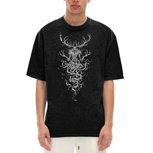 May include: A black acid-wash t-shirt featuring a white graphic print of a creature with large antlers and elongated fingers. The design extends downward in a swirling, root-like pattern, centered on the chest of the short-sleeved garment.