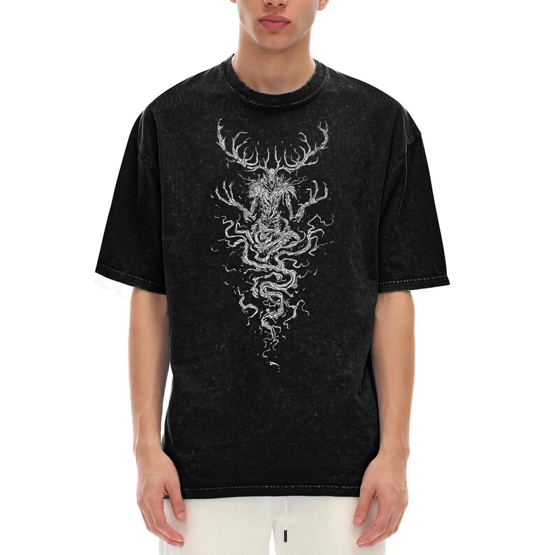 May include: A black acid-wash t-shirt featuring a white graphic print of a creature with large antlers and elongated fingers. The design extends downward in a swirling, root-like pattern, centered on the chest of the short-sleeved garment.