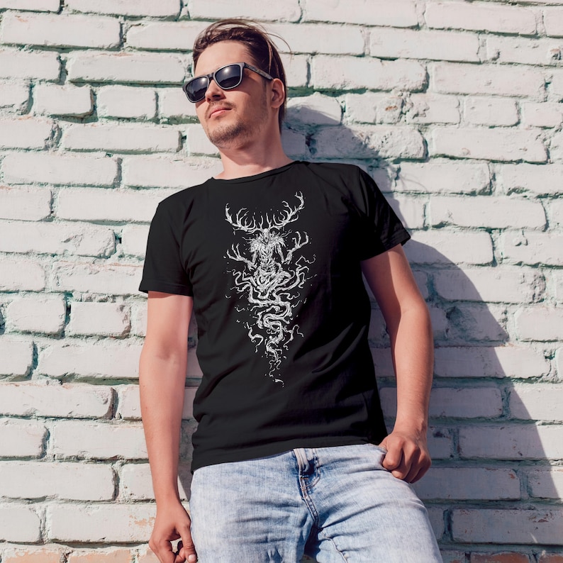 May include: A black short-sleeved t-shirt featuring a detailed white graphic print of a mythical creature with large antlers and elongated limbs, surrounded by swirling, vine-like patterns that extend downward from the center of the chest.