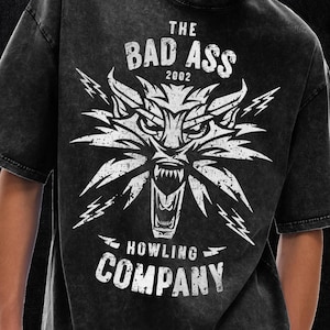 Peut inclure: Un t-shirt noir d&eacute;lav&eacute; &agrave; l'acide avec un motif blanc repr&eacute;sentant une t&ecirc;te de loup stylis&eacute;e &agrave; la gueule ouverte, entour&eacute;e d'&eacute;clairs. Le texte THE BAD ASS 2002 figure au-dessus et HOWLING COMPANY en dessous dans une police vintage us&eacute;e.