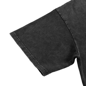 May include: A short sleeve t-shirt in a dark charcoal gray color with a washed, faded texture. The garment features a standard crew neck construction with visible stitching along the sleeve hem and the shoulder seam, showcasing a worn-in appearance.