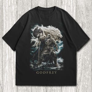 Puede incluir: Camiseta negra con un gráfico detallado de un guerrero con melena de león, sosteniendo un hacha, de pie en el agua. La palabra "GODFREY" está impresa debajo de la imagen. El diseño es en tonos grises, azules y blancos.