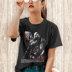 May include: A black short-sleeved t-shirt featuring a graphic print of a warrior figure with a sword. The design includes red and white accents and text that reads: An epic world tour travels across the globe and into the war-torn northern kingdoms.