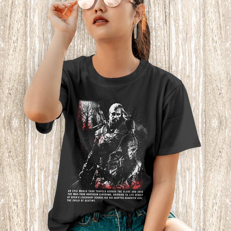 May include: A black short-sleeved t-shirt featuring a graphic print of a warrior figure with a sword. The design includes red and white accents and text that reads: An epic world tour travels across the globe and into the war-torn northern kingdoms.