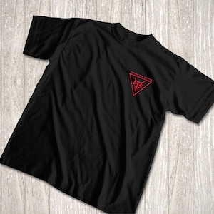 Peut inclure: T-shirt noir avec un motif rouge brodé sur le haut de la poitrine gauche. Le motif est un symbole stylisé dans un triangle rouge, avec le texte "OMNIE TIE LUPUS" au-dessus. Le t-shirt est en matière souple.