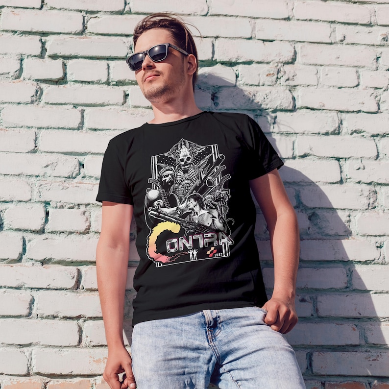 K&ouml;nnte beinhalten: Ein schwarzes Kurzarm-T-Shirt mit einem wei&szlig;en Grafikdruck des Videospiels Contra. Das Design zeigt einen Totenkopf, zwei Soldaten mit Gewehren und den Text CONTRA 1987 in einer stilisierten Schriftart mit gelb-orangem Farbverlauf.