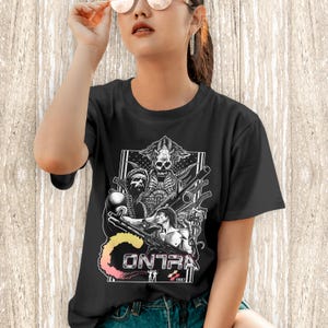 K&ouml;nnte beinhalten: Ein schwarzes Kurzarm-T-Shirt mit einem wei&szlig;en Grafikdruck des Videospiels Contra aus dem Jahr 1987. Das Design zeigt zwei muskul&ouml;se Soldaten mit Gewehren, eine sch&auml;delartige Alien-Kreatur und das Wort CONTRA in einer stilisierten Schriftart.