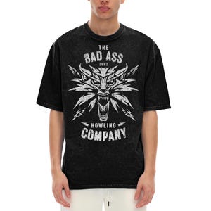 Peut inclure: Un t-shirt noir d&eacute;lav&eacute; &agrave; l'acide avec un motif blanc repr&eacute;sentant une t&ecirc;te de loup stylis&eacute;e aux crocs apparents et des &eacute;clairs. Le texte indique THE BAD ASS 2002 HOWLING COMPANY dans une police us&eacute;e. Le t-shirt a des manches courtes.