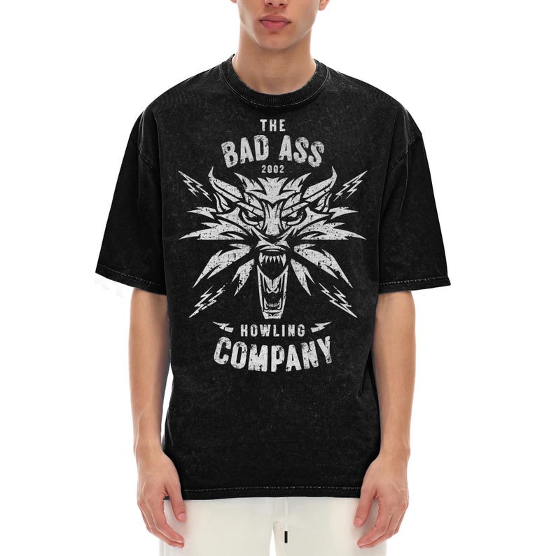 Peut inclure: Un t-shirt noir d&eacute;lav&eacute; &agrave; l'acide avec un motif blanc repr&eacute;sentant une t&ecirc;te de loup stylis&eacute;e aux crocs apparents et des &eacute;clairs. Le texte indique THE BAD ASS 2002 HOWLING COMPANY dans une police us&eacute;e. Le t-shirt a des manches courtes.