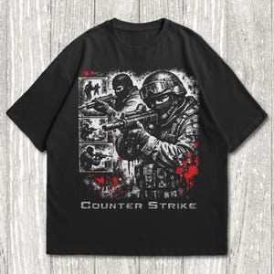 May include: Black t-shirt featuring a graphic of soldiers with weapons, and the text "Counter Strike" in white. The design includes red splatter effects and smaller images of soldiers. The shirt is a casual wear item.