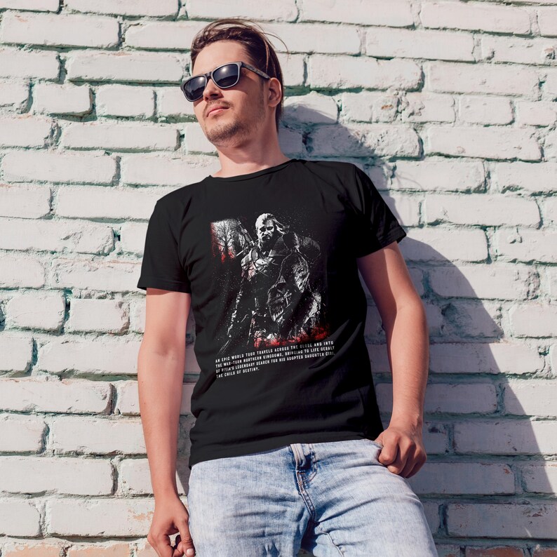 May include: A black short-sleeved t-shirt featuring a graphic illustration of a warrior with a sword and red accents. The text below the graphic reads: An epic world tour travels across the realm and into the war-torn northern kingdoms, bringing to life Geralt of Rivia's legendary search for his adopted daughter Ciri, the child of destiny.