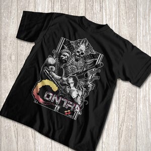K&ouml;nnte beinhalten: Ein schwarzes Kurzarm-T-Shirt mit einem Grafikdruck von zwei muskul&ouml;sen Figuren, die Gewehre halten, einer skelettartigen Alien-Kreatur und dem Text "CONTRA" mit einer bunten Rauchfahne. Das Design enth&auml;lt das Jahr 1987 und kleine Silhouetten zweier Figuren.