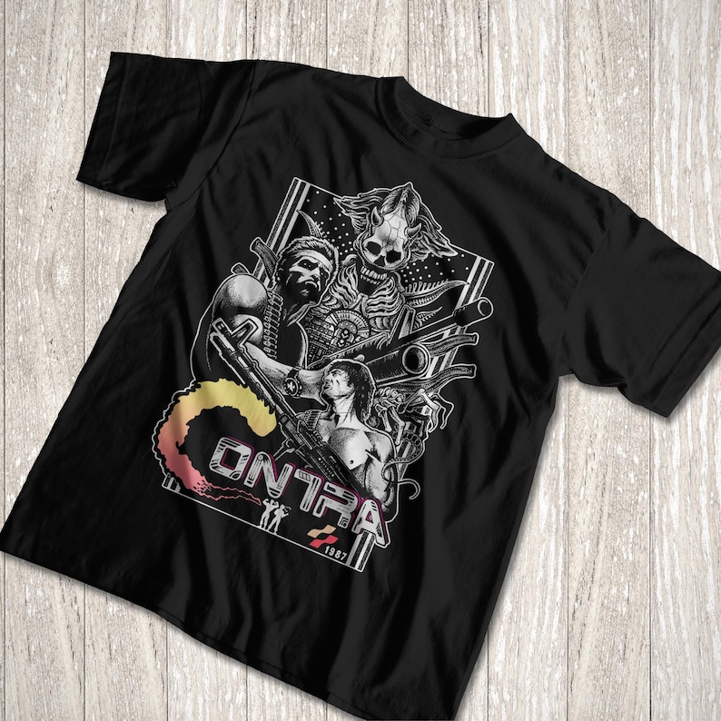 K&ouml;nnte beinhalten: Ein schwarzes Kurzarm-T-Shirt mit einem Grafikdruck von zwei muskul&ouml;sen Figuren, die Gewehre halten, einer skelettartigen Alien-Kreatur und dem Text "CONTRA" mit einer bunten Rauchfahne. Das Design enth&auml;lt das Jahr 1987 und kleine Silhouetten zweier Figuren.