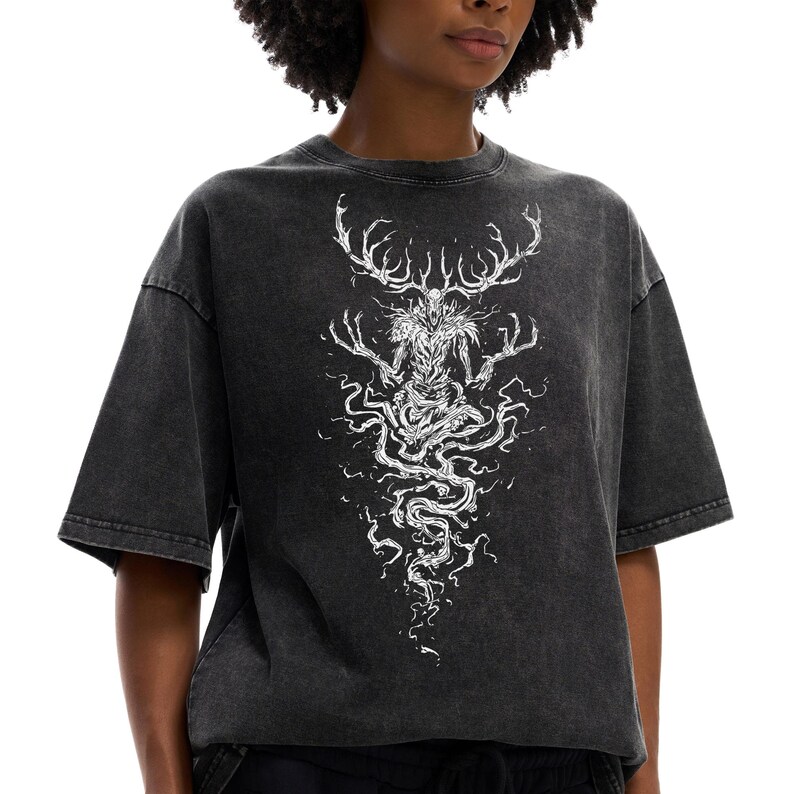 May include: A charcoal gray acid-wash t-shirt featuring a white line-art graphic of a skeletal, antlered creature with long, spindly fingers and a flowing, smoke-like lower body that tapers downward toward the hem of the garment.