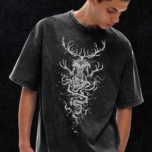 May include: A charcoal grey acid-wash t-shirt featuring a white graphic print of a skeletal creature with large antlers and long, spindly fingers. The creature is depicted in a seated position with swirling, smoke-like lines extending downward.