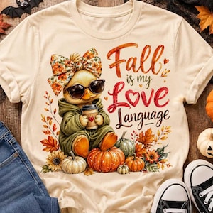 Può includere: Una t-shirt color crema con la grafica di un anatroccolo che indossa un fiocco floreale e occhiali da sole, mentre tiene una tazza di caffè. Il testo recita Fall is my Love Language. Il design include zucche, foglie autunnali e girasoli.