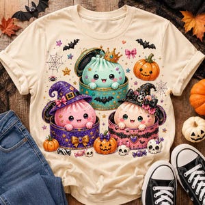 May include: A cream-colored t-shirt featuring a graphic print of three cute, round characters in open containers. One is light blue with a gold crown, one is pink with a purple witch hat, and one is light pink with a black witch hat. Includes pumpkins, bats, skulls, and spiderwebs.
