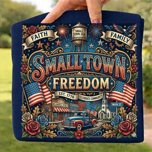 May include: A dark blue t-shirt featuring a graphic print with the text "SMALL TOWN FREEDOM" and "EST. 1776". The design includes a water tower, a vintage truck, American flags, fireworks, roses, and the words "FAITH" and "FAMILY" in banners.
