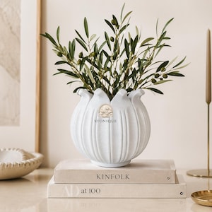 Può includere: Un vaso in ceramica bianca, strutturato, a forma di tulipano, riempito di rami di ulivo. Il vaso è appoggiato su due libri, con il testo "KINFOLK" visibile. Il vaso ha il testo "STONIQUE" sul davanti.