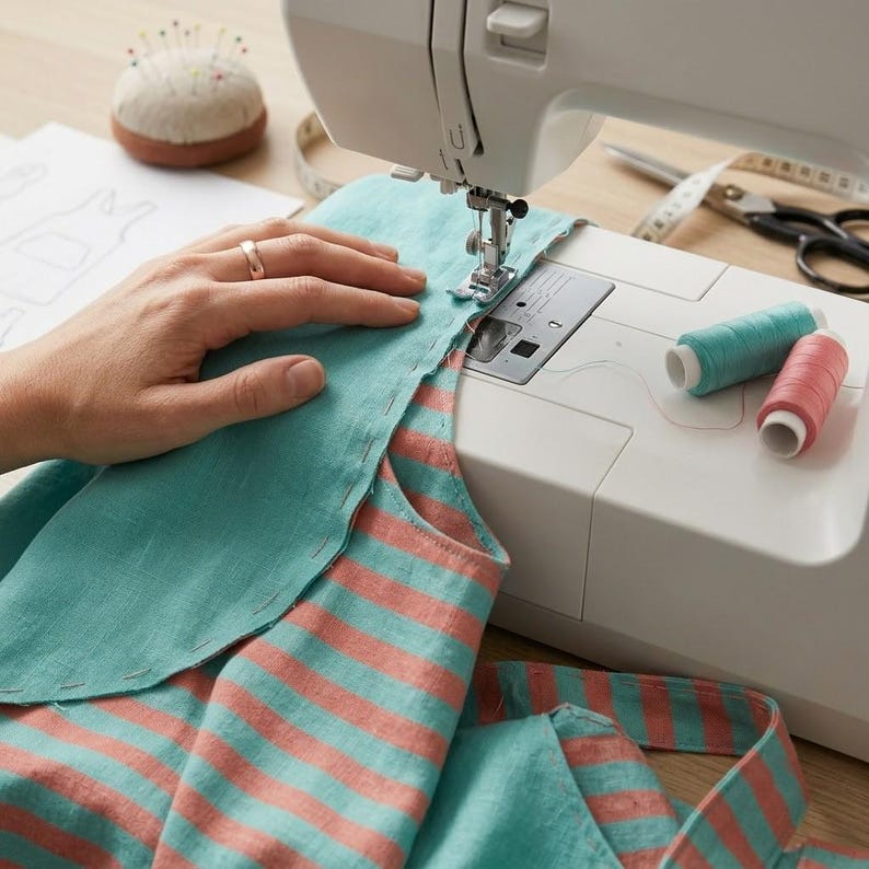 May include: A white sewing machine stitching a piece of teal and coral striped fabric. The fabric features a solid teal panel being attached to the striped section. Two spools of thread, one teal and one coral, sit on the machine base.