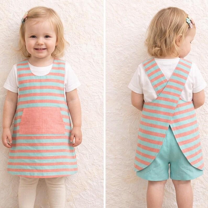 May include: A sleeveless jumper dress featuring horizontal coral and light blue stripes with a solid coral front pocket, worn over a plain white t-shirt. The back view shows a crossover strap design paired with matching light blue shorts.